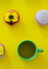 Rows of muffins decorated with powdered sugar and yellow flower shaped icing on a yellow background next to a cup of tea