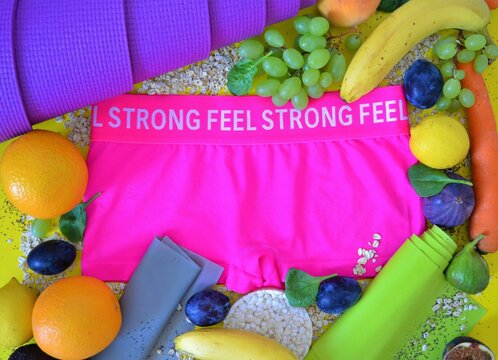Pink sweatpants with the positive message "Feel strong" surrounded by fruits, vegetables, a yoga mat and elastic bands representing a life with healthy diet and exercise or the attempt to lose weight