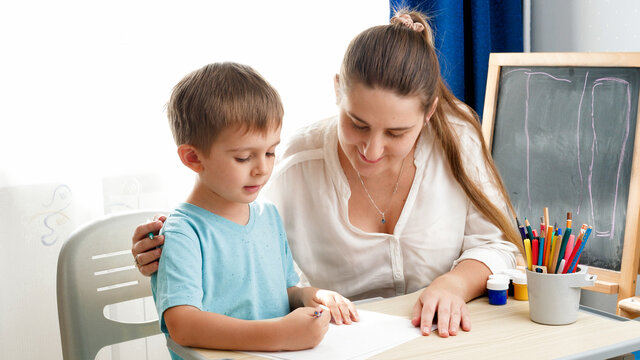 Teacher teaching drawing her little toddler son with colorful pencils at school classroom. Concept of parenting and education at home. Creativity and children art.