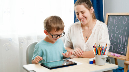 Little boy in eyeglasses playing on tablet computer. Mother hugging her little son doing homework using gadet. Children having problems with eyes and sight