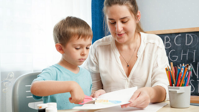 Young female teacher showing her little student how to use scissors. Boy cutting paper with scissors while doing homework.