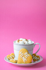 Sugar biscuits with Be Mine phrase on plate near cup of hot drink full of marshmallow on pastel pink background. Festive coffee serving or Valentines Day greeting card.