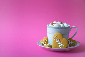 Cup of hot chocolate with marshmallow and cookie in heart shape with Love phrase on pink background. Festive coffee serving for romantic breakfast on Valentines Day. Greeting card with copy space.