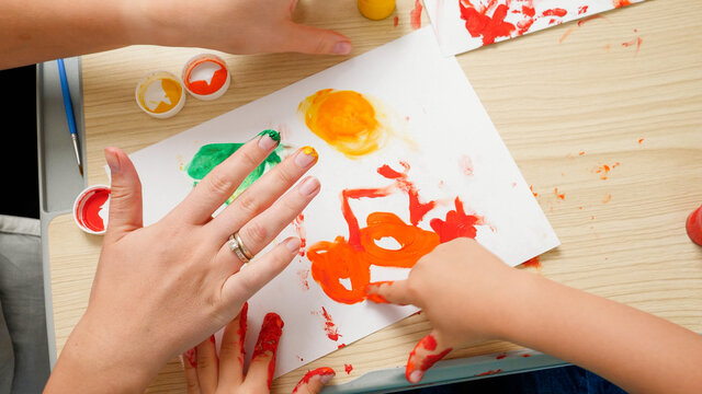 Top view of little boy with mother drawing and painting with hands covered in colorful paint on white paper. Concept of child education, creativity development and art.