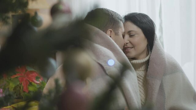 Romantic Caucasian couple sitting at Christmas tree wrapped in blanket rubbing noses. Happy husband and wife or girlfriend and boyfriend celebrating New Year at home indoors.