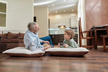 Grandfather and his cute little grandson laying together on the floor and discussing something, playing video games and spending funny time at home