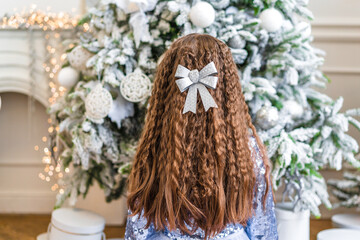 Rear view of a little girl with curly hair sits on the floor next to a Christmas tree and reaches for Christmas tree toys