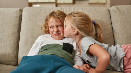 Portrait of little boy looking disgruntled while his sibling sister kissing him on cheek, sitting together on a sofa at home