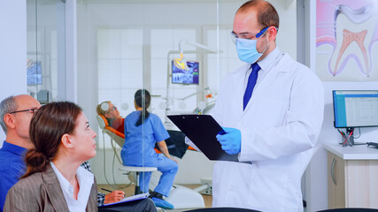 Doctor taking notes on clipboard about patient dental problems sitting on chair in waiting room of stomatological clinic. Assistant preparing old woman for examination in background