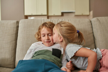 Obraz premium Gentle kiss. Portrait of cute little girl kissing her sibling brother while spending time together, cuddling on a sofa at home