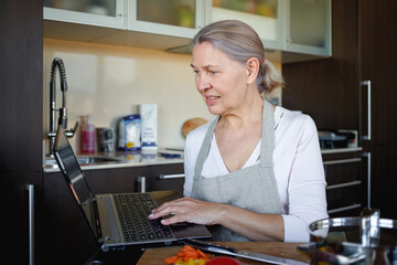 Senior woman cooking with help of recipe on laptop.