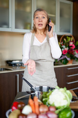 Obraz na płótnie Canvas Mature woman in kitchen preparing food and holding smart phone.