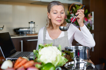 Mature woman in the kitchen prepares food and uses laptop.