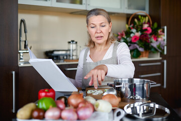 Mature woman preparing food in kitchen and looking at recipe book