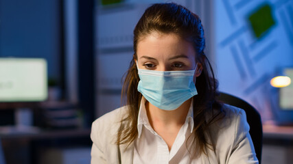 Close up of woman with face mask reading emails late at night to respect deadline of project working in new normal business office, taking notes, analysing documents overtime during global pandemic
