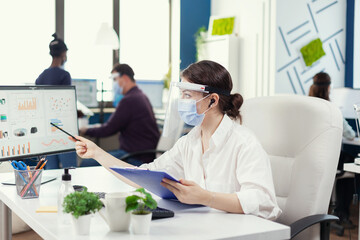 Businesswoman looking at financial stats wearing face mask during covid-19. Employees with visors working in corporate company workspace respecting social distance analysing data and graphs.