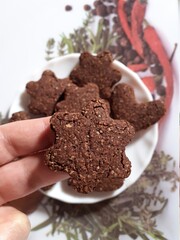 Flower shaped chocolate oat cookies, held by two fingers, with a white plate of flower and heart shaped cookies on the background.
