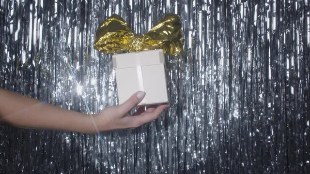 Silver tinsel rain moving and shining on the background. A hand with a gift box with a big golden bow appears and dissapears in the spotlight. 4k high quality still shot video footage.