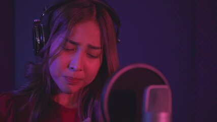 Asian young woman standing and rehearsal singing in studio with over colorful lights background. Happy female singer while recording vocals on microphone in music studio.  