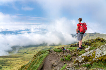A female hiker and their dog at the viewpoint from the summit of Meall Garbh near a cloudy Loch Tay...