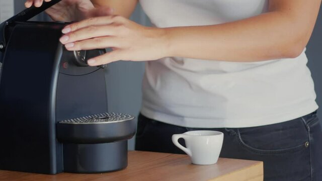 A mixed race woman in a white t-shirt putting a compostable coffee capsule into the coffee machine and making espresso in a white cup. High quality 4k video footage.