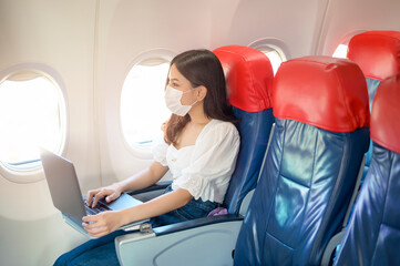 A young woman wearing face mask is using laptop onboard, New normal travel after covid-19 pandemic...