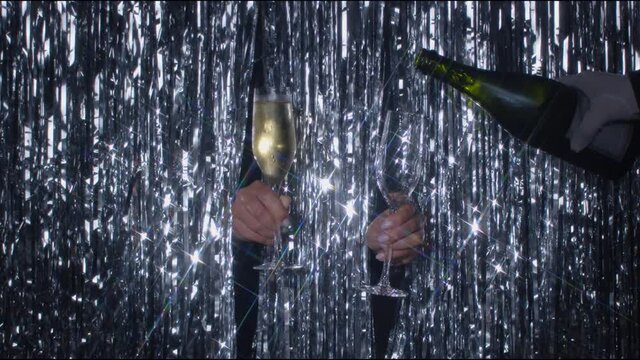 Festive silver background with two hands holding glasses with champagne. Another hand filling up one glass