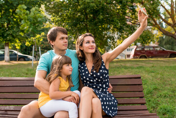 happy cheerful family in park on bench in summer, mom her daughter and dad takes pictures of themselves on a smartphone, an online application for recording video, broadcasting online to Internet.