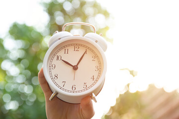 Morning time concept, hand hold white bell clock against nature sun light background .