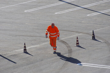 Worker in orange overalls with reflective band walks in a car-free parking lot with red and white...