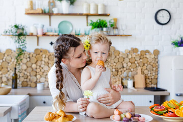Family portrait of a happy mother and daughter posing in the kitchen during breakfast, eating a croissant, cakes, cookies and fruit.