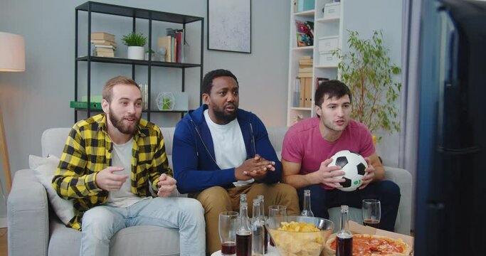 Joyful happy young African American and Caucasian men friends together watching match on TV at home. Guys screaming, hugging and celebrating goal or victory cheering fav team. football fans concept