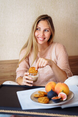 Efficient young millennial attractive woman sitting on a bed in the morning. Hand holding a cup of coffee and eating cookies. Coffee morning concept. Pleasant time during calm morning