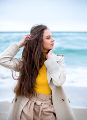 elegant brunette female with blue eyes in the coat on the winter windy beach, turquoise sea waves at the background