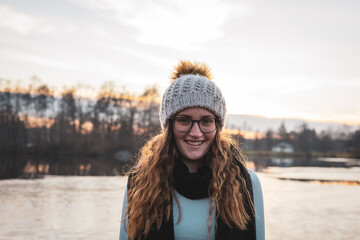Lächelnde junge Frau mit einer Wollmütze und Brille an einem See. Portrait, langes lockiges Haar, Winter.