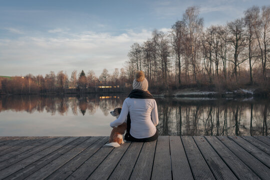 Junge Frau sitzt mit einem kleinen Terrier Hund auf einem Holzsteg am See. Rückansicht, Winter, Kälte.