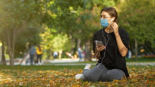 Young girl in medical protective mask sits on lawn in autumn city park and listen to music, then she takes off and throws away the mask and continues to use smartphone without it
