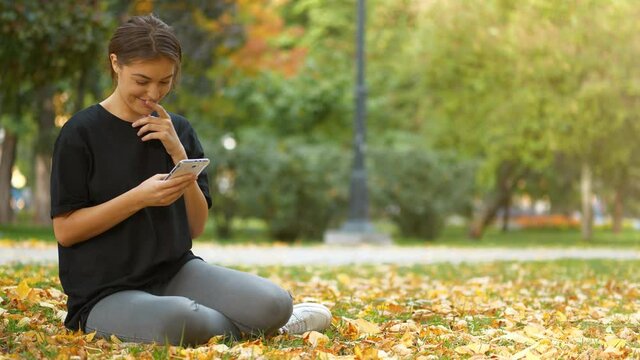 Slow Motion Young beautiful girl sits on lawn in autumn city park, looks and flips through the photos in his smartphone, she smiles, laughs and she is in a good mood