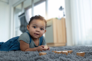 Fototapeta premium Little dark skinned girl lay on the carpet at home, and playing wooden toys at home, looking at camera