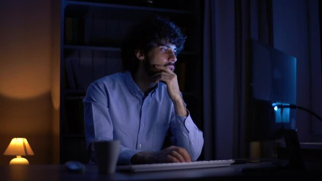 Thoughtful young businessman working on computer at late night at home office. Focused freelancer looking screen in dark room at evening. Overworked male using pc at apartment.