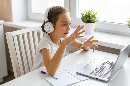 A girl student in headphones in front of a laptop monitor talk with a teacher during an online lesson.