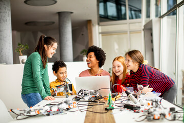 Happy kids with their African American female science teacher with laptop programming electric toys...