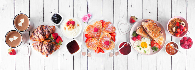 Valentines or Mothers Day breakfast table scene. Above view on a white wood banner background. Heart shaped pancakes, eggs and a variety of love themed food.