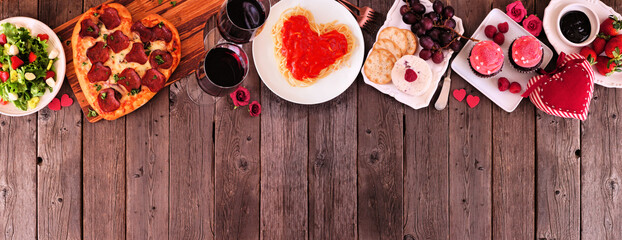 Fototapeta na wymiar Home cooked Valentines Day dinner. Overhead view top border against a dark wood banner background. Heart shaped pizza, pasta, wine, cheese plate and desserts.