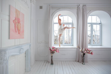 Ballerina poses in front of a large window, standing on the windowsill. Girl in a white ballet skirt.