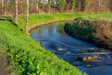 Small winding river with a calm flow between stones among green grass and trees, sunny winter day...