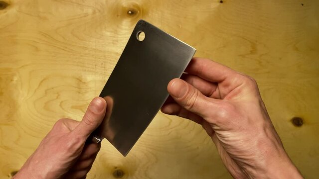 A man holds a kitchen cleaver and checks its edge for sharpness. Chromed cleaver on a wooden background. Close-up. Top view.