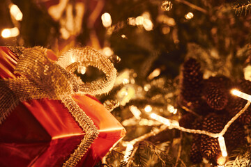 Horizontal photography of big red present with golden rustic style bow on Christmas Fir Tree with garland lights