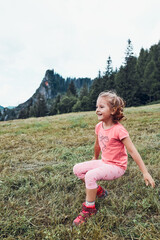 Little girl laying playing on grass enjoying summer day. Happy child playing in the field during vacation trip in mountains. Mountain landscape in the background