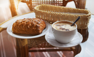 A cup of coffee with a fresh bun on a glass table in a cafe in the early morning. Selective focus. Soft light. Comfort.